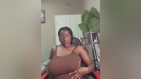 Media: A video shows a Black woman with braids, wearing a brown tank top, seated in a red chair, with a green plant and shelves in the background.