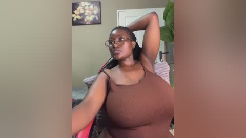 Media: A video of a curvy Black woman with dark skin and long hair, wearing a brown ribbed tank top, posing confidently in a bedroom with beige walls and a flower painting.