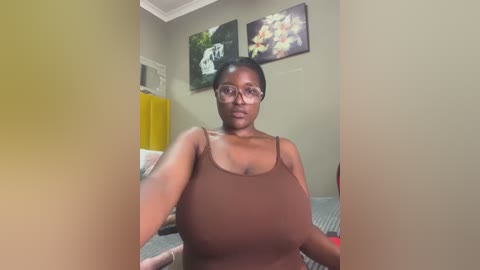 Media: Video of a dark-skinned woman with glasses, medium build, wearing a brown tank top, standing in a beige room with framed art on the wall and a yellow dresser.