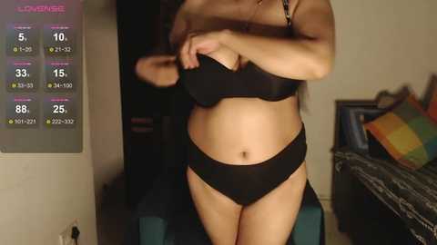 Media: Video of a light-skinned woman with long hair, wearing black lingerie, lifting her bra, in a dimly-lit bedroom with a digital health monitor on the wall.