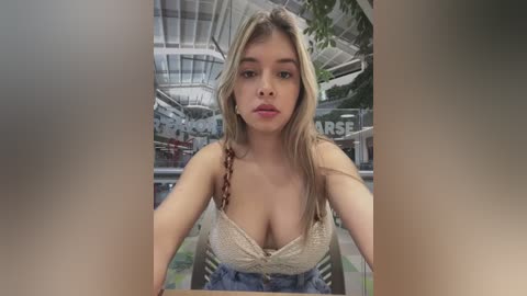 Video of a young Caucasian woman with long blonde hair, wearing a low-cut, beige lace top and high-waisted blue jeans, standing in a modern, brightly lit indoor mall.