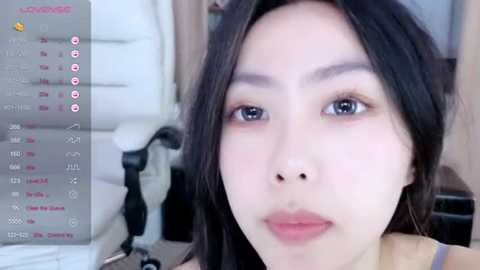 Video of an Asian woman with long black hair, fair skin, and light makeup, sitting in an office chair, taken from a webcam.