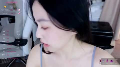 A video of a young East Asian woman with fair skin and short black hair, wearing a light blue spaghetti strap top. She appears contemplative, indoors, with a blurred background of a white chair and beige curtain.