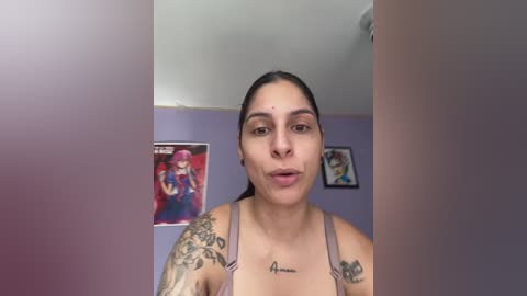 Video of a Latina woman with medium brown skin and dark hair, wearing a pink tank top, standing in a room with purple walls, featuring two framed anime-style posters.