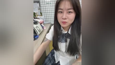 Media: A video of a young Asian woman with long black hair, wearing a white shirt, plaid skirt, and tie, sitting at a desk in an office. The background includes office supplies and a whiteboard.