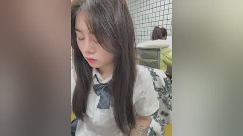 Media: A video of a young Asian woman with long black hair, wearing a white blouse with a blue bow tie, sitting in a public restroom with a tiled wall and green patterned floor.