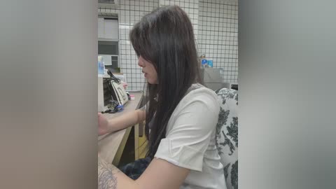 Media: Video of a young woman with long brown hair, sitting at a wooden desk in a cluttered office, wearing a white t-shirt with a floral design.