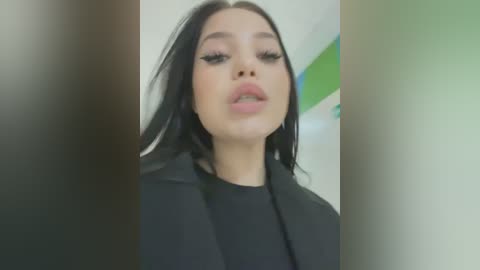 Video of a young woman with fair skin and long black hair, wearing black lipstick and a black blazer, captured in a blurred, out-of-focus setting.