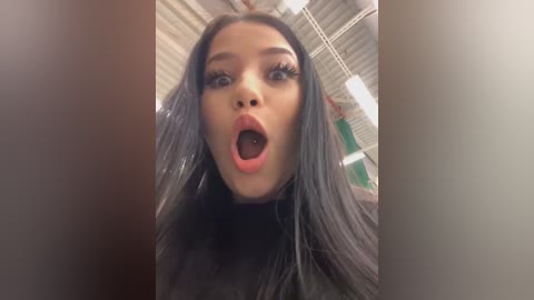 Video of a young Asian woman with long, straight black hair, wearing a black turtleneck, mouth open in surprise, standing in an industrial building with exposed pipes and fluorescent lights.