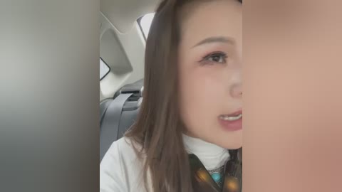 Media: Video of a young Asian woman with long brown hair, wearing a white turtleneck and a colorful scarf, seated in a car with gray seats and a visible sunroof.