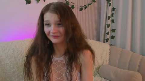 Video of a young Caucasian woman with long brown hair and fair skin, wearing a sheer, white lace top, smiling softly. She's in a cozy room with a white couch, green ivy, and purple lighting.