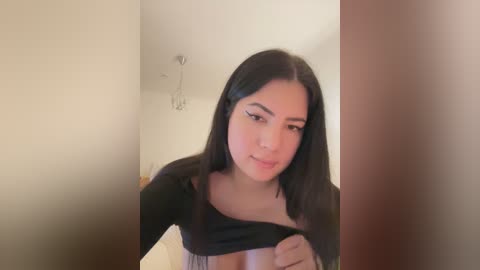 Media: Video of a young Latina woman with straight black hair, light skin, and dark eyebrows, lifting her black top to reveal cleavage. Background shows beige walls and a metallic wall hook.