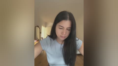 Media: Video of a young woman with long black hair, wearing a light blue T-shirt, looking down, standing in a dimly lit, beige-walled room with a wooden dresser in the background.