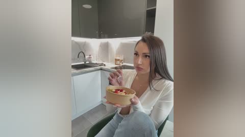 Video of a young woman with long brown hair, light skin, and medium build, wearing a white top, eating a yogurt bowl in a modern kitchen with white cabinets and grey tiles.