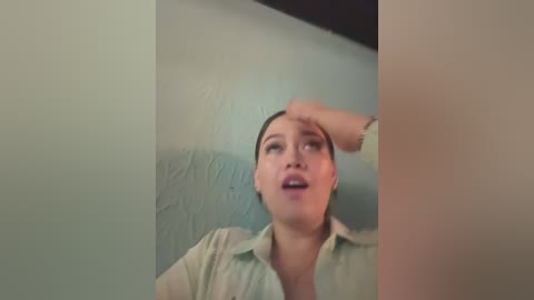 A video of a young woman with fair skin, dark hair pulled back, wearing a light green button-up shirt, leaning against a textured teal wall, her mouth slightly open, hand resting on her forehead, suggesting a moment of thought or frustration.