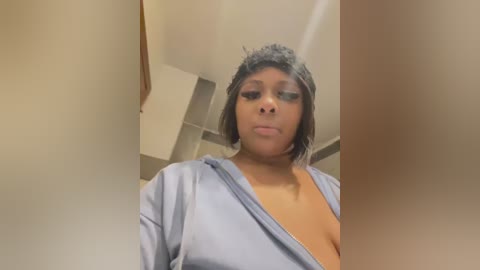 A video of a Black woman with short, curly gray hair and a light blue robe, partially open, revealing her breasts, taken in a bathroom with beige tiles and a white ceiling.