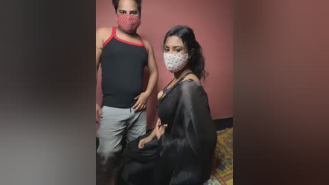 Media: Video of a man in a red face mask, black tank top, and grey pants standing next to a woman in a black face mask, black dress, and a patterned shawl, both with medium skin tones.