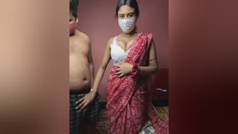 Media: Video of a young woman in a red sari with a white lace bra, holding hands with a shirtless man wearing black pants, both masked, in a dimly lit room with red walls.