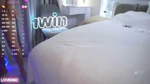 Video of a serene, neatly made bed with white sheets and a gray upholstered headboard, set in a bright, modern bedroom with a vanity table and a large window. The word \"Lovesie\" is overlaid in white text.