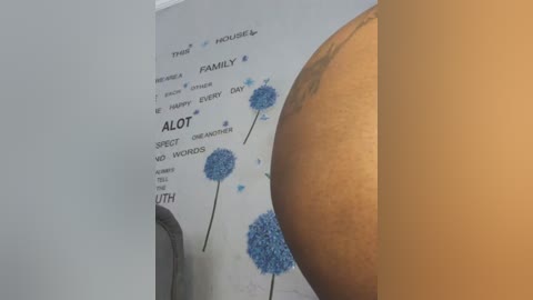 Media: Video of a wall-mounted poster featuring blue dandelion illustrations with \"ALOT\" and \"LOVE\" text, alongside a partially visible tattooed arm. Background includes a beige wall and part of a gray chair.