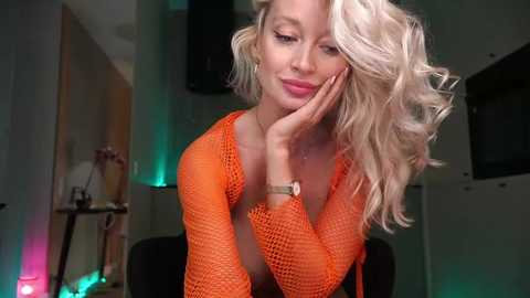 Media: Video of a blonde woman with wavy hair, wearing an orange fishnet top, leaning on her hand, smiling softly, in a dimly-lit room with green and blue lights.