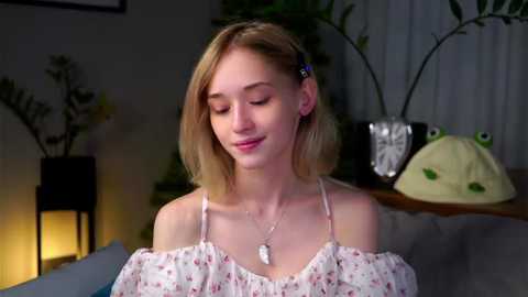 Media: Video of a fair-skinned, young woman with straight blonde hair, wearing a floral off-shoulder top, smiling softly, surrounded by potted plants and a frog lamp.