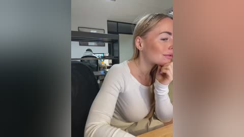 Media: Video of a blonde woman with fair skin and medium build, wearing a white long-sleeve top, sitting in a modern office with black furniture and muted tones.