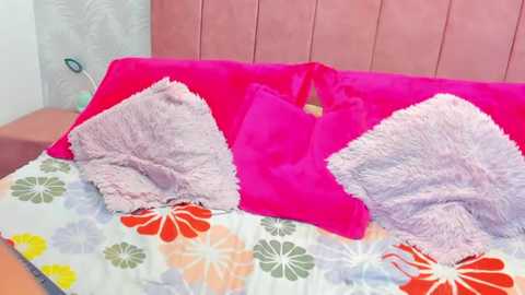 Video of a bed with a vibrant pink headboard, a fluffy white pillow, and a bright pink pillow. The bedspread features a colorful floral pattern in shades of orange, green, and white.