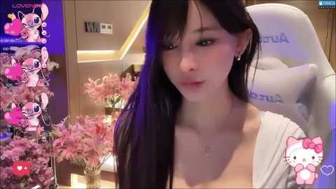 Media: A video of an Asian woman with long black hair, fair skin, and a small bust, wearing a white top, sitting in a gaming chair. The background features a pink floral arrangement and a plush My Melody toy.