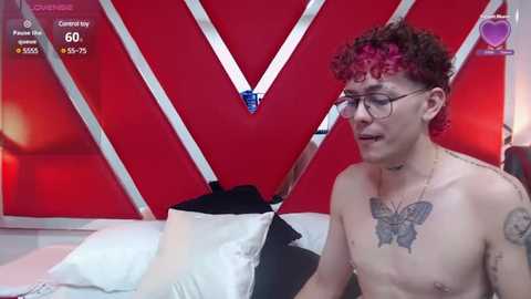 Media: Video of a shirtless young man with curly red hair, glasses, and tattoos, seated on a bed with white pillows. Background features a red and white geometric design.