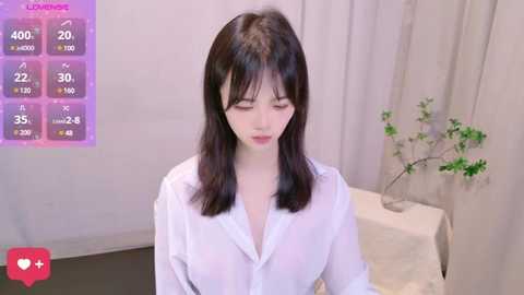 Video of an East Asian woman with straight black hair, wearing a white button-up shirt, in a minimalist room with a table, green plant, and cream curtains.