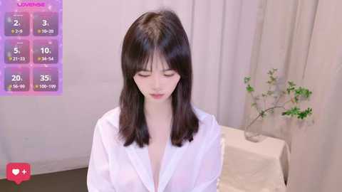 A video of a young East Asian woman with long, straight black hair, wearing a white blouse, standing in a minimalist room with a vase of greenery on a white table.