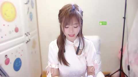 Media: Video of an Asian woman with long brown hair, wearing a white dress, seated in a brightly colored, child-themed bathroom with a green emergency light above.