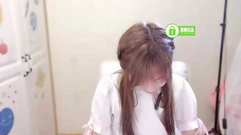 Media: Video of a young Asian woman with long brown hair in a white robe, sitting in a clean, beige bathroom with a green \"1\" sticker on her head, indicating hair dye application.