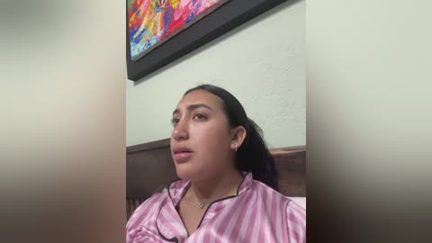 Video of a young woman with light brown skin, dark hair, and medium build, wearing a pink and white striped shirt, sitting indoors with a colorful abstract painting on the wall behind her.