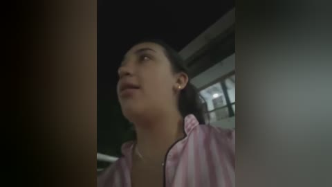 Video of a young woman with fair skin and dark hair, wearing a light pink jacket, looking upward, indoors, with dim lighting.