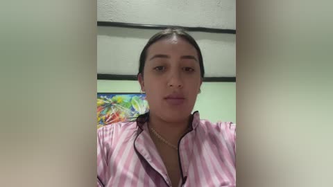 Video of a young woman with medium-brown skin, dark hair pulled back, wearing a pink and white striped blouse, standing indoors with a colorful painting in the background.