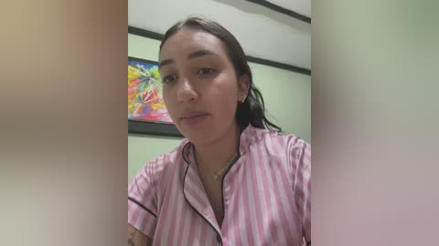 A video of a young Latina woman with light skin and dark hair, wearing a pink and white striped blouse, standing in front of a colorful painting in a green-walled room.