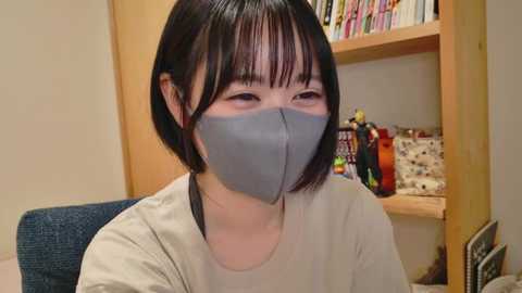 Video of an Asian woman with short black hair, wearing a gray face mask, light gray t-shirt, and sitting in a room with a wooden shelf filled with books and a red plastic bottle.