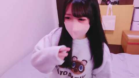 Video of an East Asian woman with long black hair, wearing a white mask, white hoodie, and a \"Kitty\" graphic tee, holding a phone, indoors.