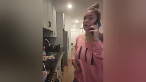 Video of a woman in a pink sweater talking on her phone in a modern kitchen with white cabinets and wooden floors.