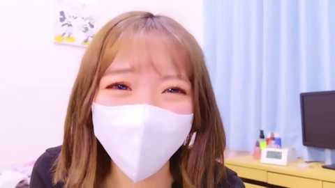 Media: Video of an Asian woman with light brown hair, wearing a white surgical mask, in a modern office with light blue curtains and a desk with office supplies.