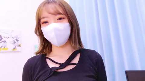 Media: Video of an East Asian woman with light skin, shoulder-length straight brown hair, wearing a white face mask and a black top with a cut-out design. Background includes a white wall with a black and white artwork and a blue curtain.