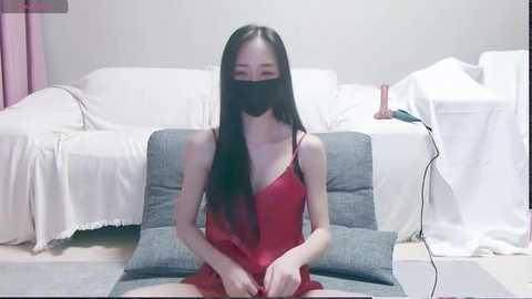 Video of an East Asian woman with long black hair, fair skin, and a slender physique, wearing a red nightgown and mask, seated on a grey cushion, in a minimalist room with white furniture.