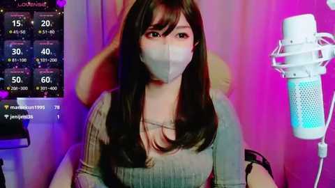 Media: Video of an Asian woman with long black hair wearing a face mask, seated in a chair. Background shows a colorful, illuminated screen displaying temperature and humidity data.