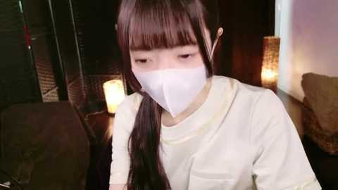 Media: Video of an Asian woman with long brown hair, wearing a white face mask and a white T-shirt, sitting indoors, dimly lit with a wicker lamp and dark furniture in the background.