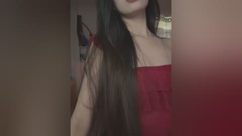 Media: Video of a young Asian woman with long black hair, wearing a red off-shoulder top, captured in a dimly lit room with blurred background objects.