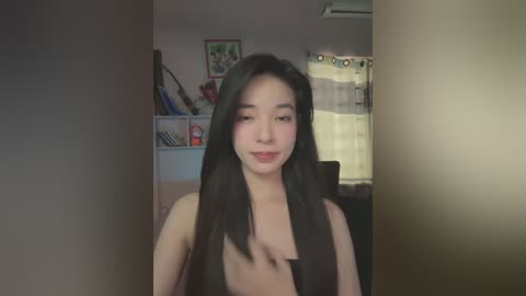 Media: Video of an Asian woman with long black hair, wearing a black top, standing in a dimly lit room with white curtains and bookshelves in the background.