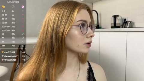 Video of a fair-skinned, red-haired young woman with glasses, wearing a black lace top, in a modern kitchen with white cabinets and a coffee machine.