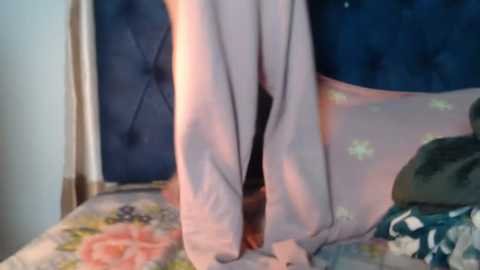 A video of a person wearing a light pink shirt and white underwear, standing on a bed with floral-patterned sheets and a dark blue quilted headboard in the background.